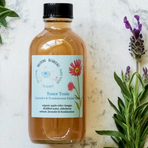May include: A clear glass bottle with a black cap filled with a light brown liquid. The bottle has a white label with the text "BALLERINA BOTANICALS Toner Tonic Lavender & Frankincense Facial Toner organic apple cider vinegar, distilled water, elderberry extract, lavender & frankincense".