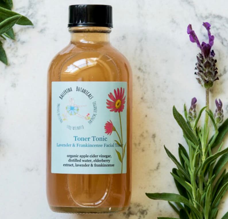 May include: A clear glass bottle with a black cap filled with a light brown liquid. The bottle has a white label with the text "BALLERINA BOTANICALS Toner Tonic Lavender & Frankincense Facial Toner organic apple cider vinegar, distilled water, elderberry extract, lavender & frankincense".