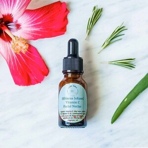 May include: A brown glass bottle with a dropper top containing Hibiscus Infused Vitamin C Facial Nectar. The bottle is labeled with the product name and ingredients, including aloe vera, jojoba oil, argan oil, hibiscus petals, patchouli oil, rosehip oil, and rosemary oil.