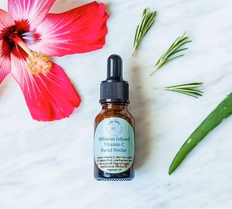 May include: A brown glass bottle with a dropper top containing Hibiscus Infused Vitamin C Facial Nectar. The bottle is labeled with the product name and ingredients, including aloe vera, jojoba oil, argan oil, hibiscus petals, patchouli oil, rosehip oil, and rosemary oil.
