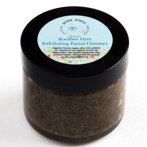 May include: A black jar with a blue and white label that reads "Rooibos Mint Exfoliating Facial Cleanser". The label lists the ingredients: organic brown sugar, aloe vera, jojoba oil, vitamin E oil, apricot kernel oil, shea butter, raw honey, bentonite clay, rooibos leaves, chamomile oil, lemon oil, and peppermint oil.
