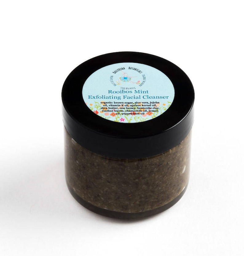 May include: A glass jar with a black lid containing a brown exfoliating facial cleanser. The label on the jar reads "Rooibos Mint Exfoliating Facial Cleanser" and lists the ingredients: organic brown sugar, aloe vera, jojoba oil, vitamin E oil, apricot kernel oil, shea butter, raw honey, bentonite clay, rooibos leaves, chamomile oil, lemon oil, peppermint oil.