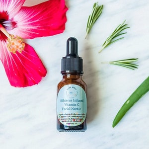 May include: A small brown glass bottle with a dropper top. The bottle is labeled "Hibiscus Infused Vitamin C Facial Nectar" and lists ingredients including vitamin C, aloe vera, jojoba oil, vitamin E oil, meadowfoam seed oil, argan oil, hibiscus petals, patchouli oil, frankincense oil, and rosemary oil.