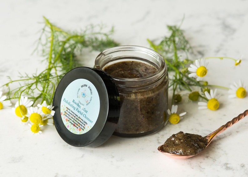 May include: A glass jar of dark green facial cleanser with a black lid. The lid has the text "Rooibos Mint Exfoliating Facial Cleanser" printed on it. The jar is sitting on a white marble surface with a small spoon of the cleanser next to it. There are sprigs of chamomile flowers in the background.