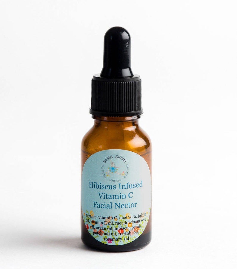 May include: A brown glass bottle with a black dropper top. The bottle has a white label with the text "Hibiscus Infused Vitamin C Facial Nectar" and a list of ingredients.
