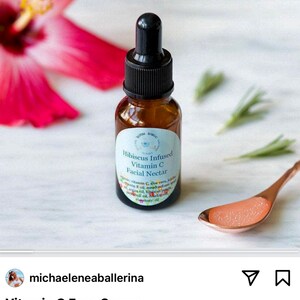 May include: A small amber glass bottle with a dropper top, labeled "Hibiscus Infused Vitamin C Facial Nectar". The bottle is sitting on a white surface with a pink hibiscus flower and green sprigs in the background. A small gold spoon with a clear liquid is also visible.