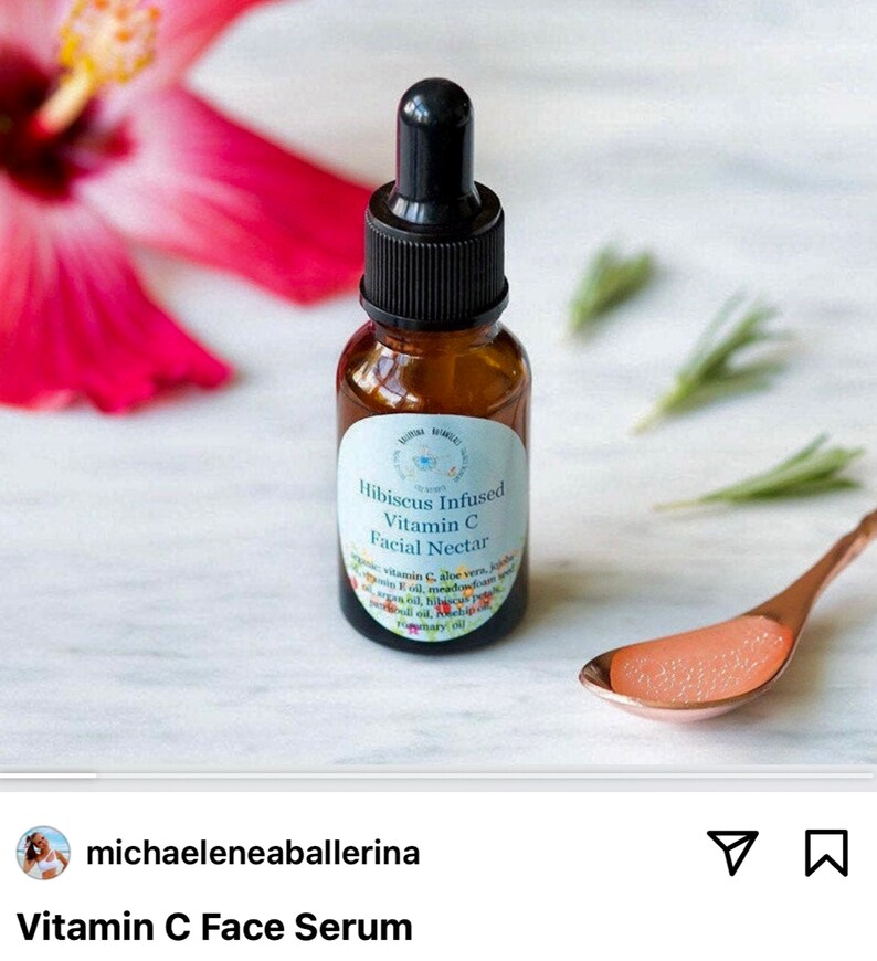 May include: A small amber glass bottle with a dropper top, labeled "Hibiscus Infused Vitamin C Facial Nectar". The bottle is sitting on a white surface with a pink hibiscus flower and green sprigs in the background. A small gold spoon with a clear liquid is also visible.
