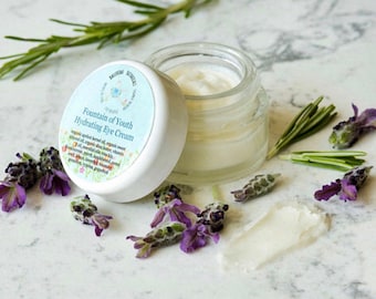 Può includere: Un barattolo bianco con un coperchio bianco contenente una crema bianca. Il barattolo ha un'etichetta con la scritta "Fountain of Youth Hydrating Eye Cream". Il barattolo è circondato da fiori di lavanda e rametti di rosmarino su una superficie di marmo bianco.