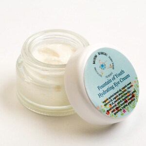 May include: A small glass jar with a white lid containing a white cream. The lid has a blue and white label with the text "Fountain of Youth Hydrating Eye Cream" and a list of ingredients.