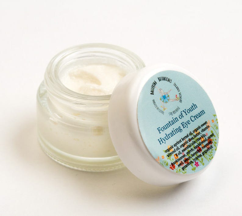 May include: A small glass jar with a white lid containing a white cream. The lid has a blue and white label with the text "Fountain of Youth Hydrating Eye Cream" and a list of ingredients.