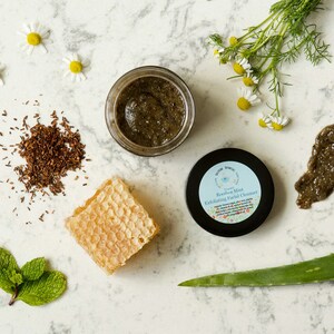 May include: A small jar of green Rooibos Mint Exfoliating Facial Cleanser with a white label and a blue border. The jar is surrounded by ingredients including chamomile flowers, dried rooibos tea, honeycomb, aloe vera, and fresh mint leaves.
