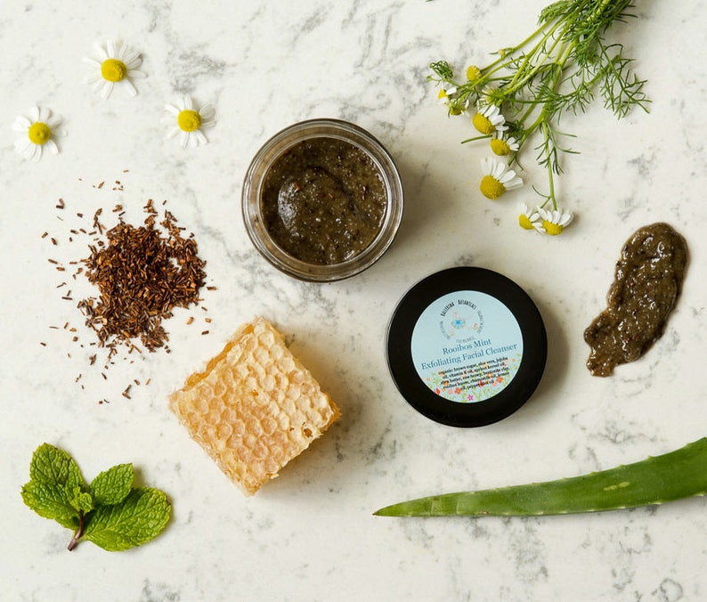 May include: A small jar of green Rooibos Mint Exfoliating Facial Cleanser with a white label and a blue border. The jar is surrounded by ingredients including chamomile flowers, dried rooibos tea, honeycomb, aloe vera, and fresh mint leaves.