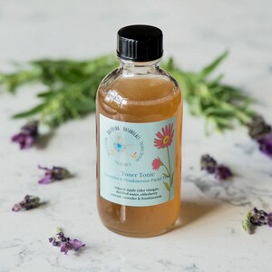 May include: A clear glass bottle with a black lid filled with a light brown liquid. The bottle has a blue and white label with the text "Toner Tonic Lavender & Frankincense Facial Toner" and a floral design.