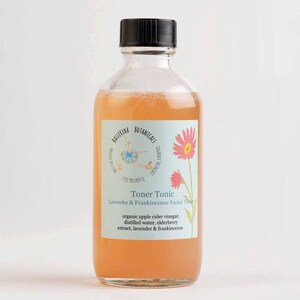 May include: A clear glass bottle with a black cap filled with a light orange liquid. The label is light blue with a pink flower and a ballerina illustration. The label reads "Ballerina Botanicals Toner Tonic Lavender & Frankincense Facial Toner organic apple cider vinegar, distilled water, elderberry extract, lavender & frankincense"
