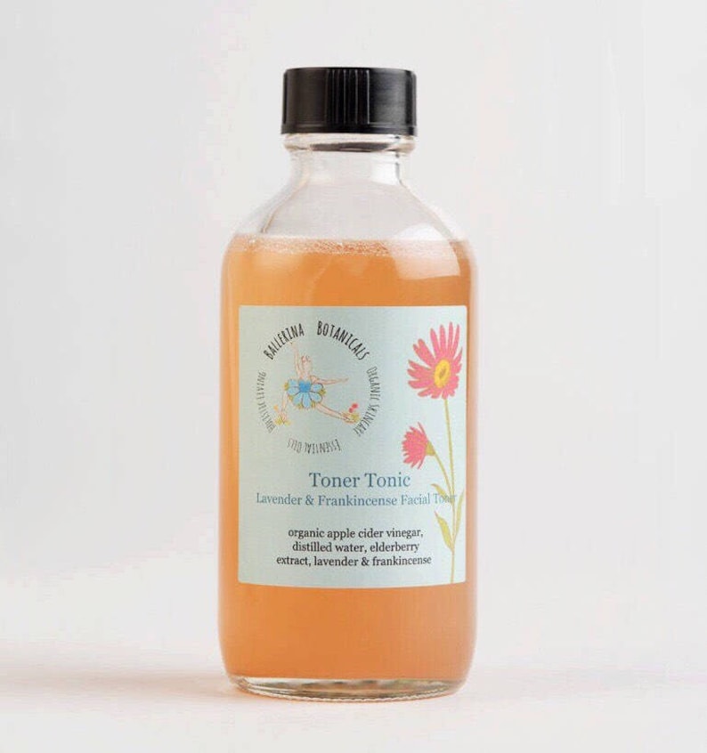 May include: A clear glass bottle with a black cap filled with a light orange liquid. The label is light blue with a pink flower and a ballerina illustration. The label reads "Ballerina Botanicals Toner Tonic Lavender & Frankincense Facial Toner organic apple cider vinegar, distilled water, elderberry extract, lavender & frankincense"