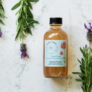 May include: A clear glass bottle with a black cap filled with a light brown liquid. The bottle is labeled "Toner Tonic Lavender & Frankincense Facial Toner" and lists the ingredients: organic apple cider vinegar, distilled water, elderberry extract, lavender & frankincense.
