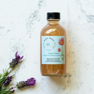 May include: A glass bottle of toner tonic with a light brown liquid. The label is blue and white with the text "Toner Tonic Lavender & Frankincense Facial Toner" and a list of ingredients: organic apple cider vinegar, distilled water, elderberry extract, lavender & frankincense.