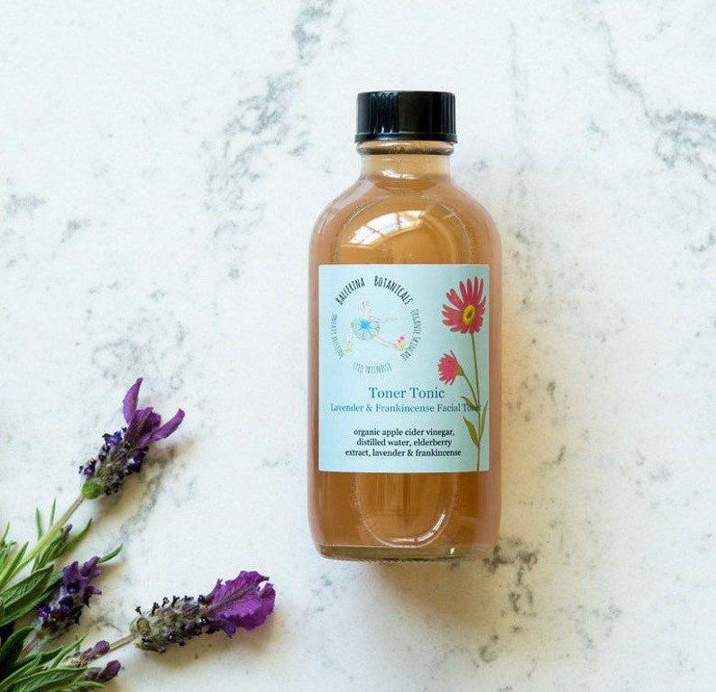 May include: A glass bottle of toner tonic with a light brown liquid. The label is blue and white with the text "Toner Tonic Lavender & Frankincense Facial Toner" and a list of ingredients: organic apple cider vinegar, distilled water, elderberry extract, lavender & frankincense.