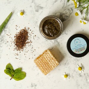 May include: A close-up of a green and brown facial scrub in a glass jar. The jar is labeled "Rooibos Mint Exfoliating Facial Cleanser". The scrub is surrounded by natural ingredients like aloe vera, chamomile flowers, and honeycomb.