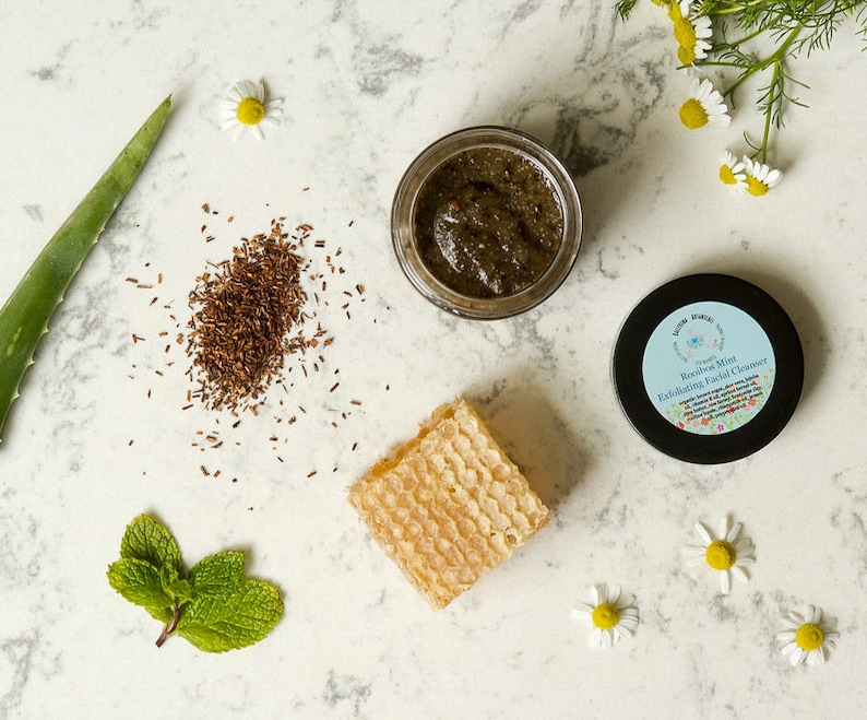 May include: A close-up of a green and brown facial scrub in a glass jar. The jar is labeled "Rooibos Mint Exfoliating Facial Cleanser". The scrub is surrounded by natural ingredients like aloe vera, chamomile flowers, and honeycomb.