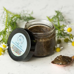 May include: A glass jar of dark brown, grainy facial cleanser with a black lid labeled "Rosemary Mint Exfoliating Facial Cleanser". The jar is sitting on a white surface with a small spoon containing the cleanser and a sprig of white chamomile flowers.