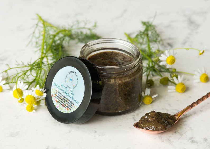 May include: A glass jar of dark brown, grainy facial cleanser with a black lid labeled "Rosemary Mint Exfoliating Facial Cleanser". The jar is sitting on a white surface with a small spoon containing the cleanser and a sprig of white chamomile flowers.