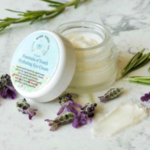 May include: A white jar with a blue and green label that reads "Fountain of Youth Hydrating Eye Cream". The jar is open and the cream is visible. The jar is surrounded by lavender flowers and rosemary sprigs.
