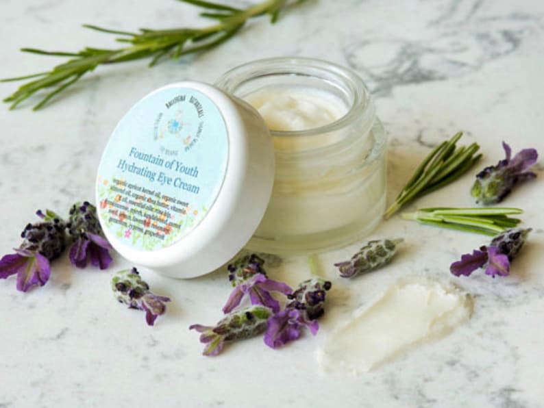 May include: A white jar with a blue and green label that reads "Fountain of Youth Hydrating Eye Cream". The jar is open and the cream is visible. The jar is surrounded by lavender flowers and rosemary sprigs.