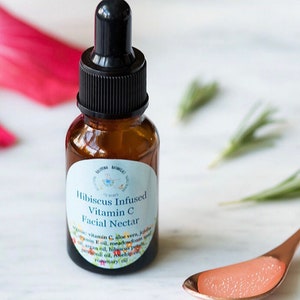 May include: A brown glass bottle with a black dropper top and a white label that reads "Hibiscus Infused Vitamin C Facial Nectar". The label lists ingredients including aloe vera, jojoba, vitamin E oil, meadowfoam seed oil, argan oil, hibiscus petals, patchouli oil, and rosemary oil.