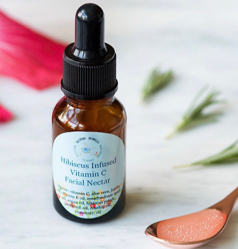 May include: A brown glass bottle with a black dropper top and a white label that reads "Hibiscus Infused Vitamin C Facial Nectar". The label lists ingredients including aloe vera, jojoba, vitamin E oil, meadowfoam seed oil, argan oil, hibiscus petals, patchouli oil, and rosemary oil.