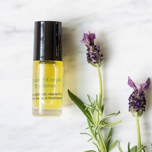 May include: A clear glass bottle with a black cap and yellow liquid inside. The bottle is labeled "Vitamin E Complex Eye Serum" and lists ingredients including jojoba oil, vitamin E oil, lavender, lemon, and frankincense. The bottle is sitting on a white marble surface with lavender flowers.