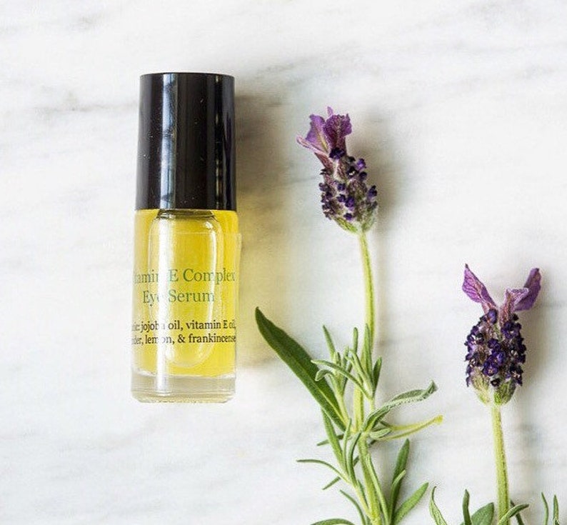 May include: A clear glass bottle with a black cap and yellow liquid inside. The bottle is labeled "Vitamin E Complex Eye Serum" and lists ingredients including jojoba oil, vitamin E oil, lavender, lemon, and frankincense. The bottle is sitting on a white marble surface with lavender flowers.