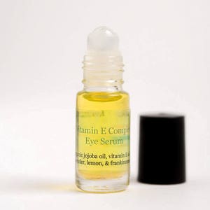 May include: A clear glass bottle with a black cap, containing a yellow liquid. The bottle is labeled "Vitamin E Complex Eye Serum" and lists ingredients including jojoba oil, vitamin E, lavender, lemon, and frankincense.