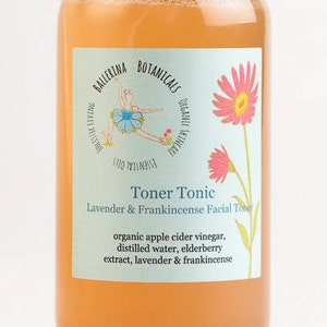 May include: A clear glass bottle with a light orange liquid inside. The label is light blue with pink flowers and a ballerina illustration. The label reads "Toner Tonic Lavender & Frankincense Facial Toner" and lists the ingredients: "organic apple cider vinegar, distilled water, elderberry extract, lavender & frankincense".