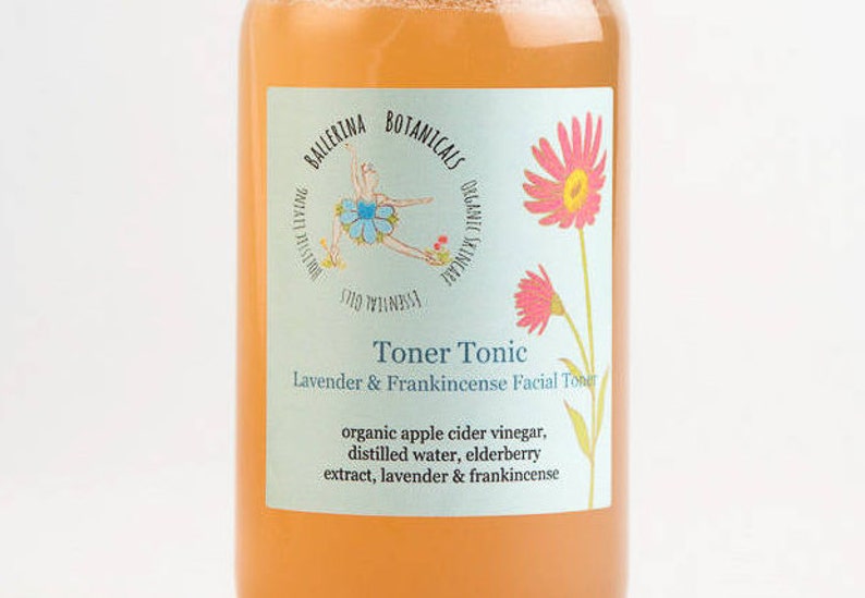 May include: A clear glass bottle with a light orange liquid inside. The label is light blue with pink flowers and a ballerina illustration. The label reads "Toner Tonic Lavender & Frankincense Facial Toner" and lists the ingredients: "organic apple cider vinegar, distilled water, elderberry extract, lavender & frankincense".