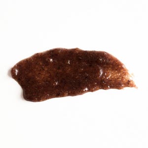 May include: A brown, grainy, and slightly textured substance spread on a white surface. The substance appears to be a facial scrub or mask.