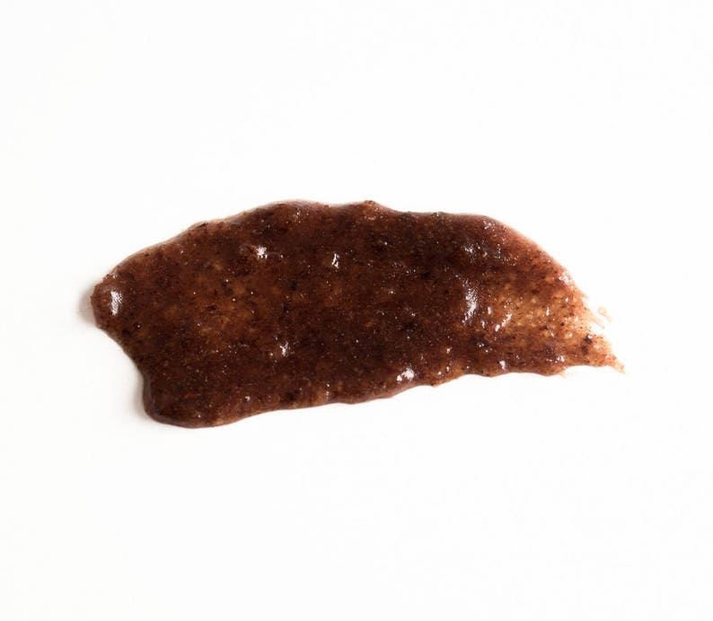 May include: A brown, grainy, and slightly textured substance spread on a white surface. The substance appears to be a facial scrub or mask.
