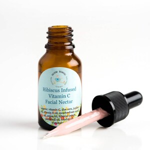 May include: A small amber glass bottle with a dropper filled with a pink liquid. The bottle has a white label with the text "Hibiscus Infused Vitamin C Facial Nectar" and a list of ingredients.