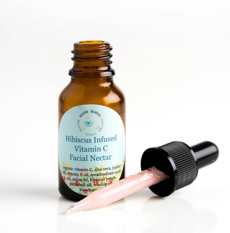 May include: A small amber glass bottle with a dropper filled with a pink liquid. The bottle has a white label with the text "Hibiscus Infused Vitamin C Facial Nectar" and a list of ingredients.