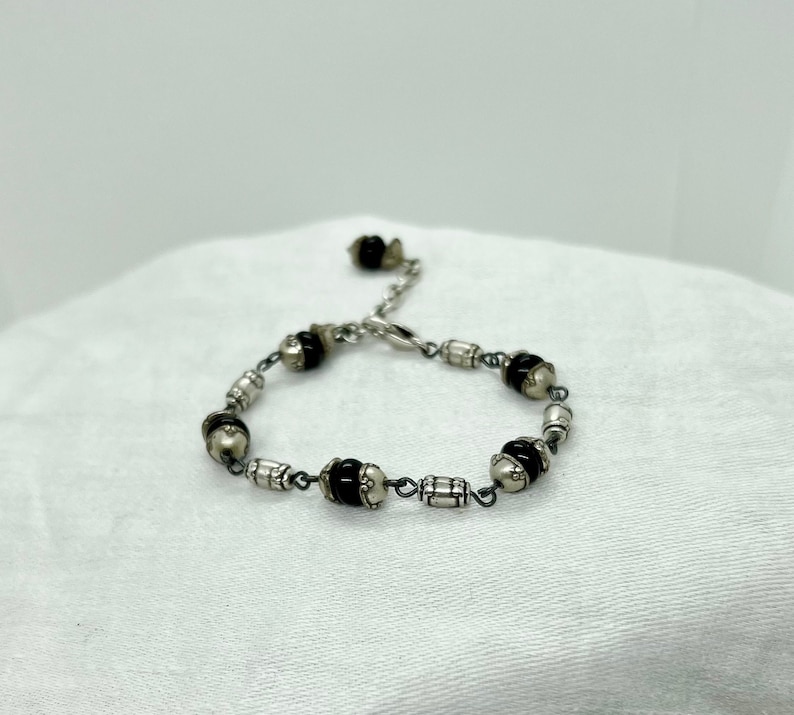 Puede incluir: Un brazalete de plata con cuentas negras y blancas. El brazalete tiene un cierre de broche.