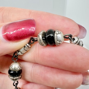 Puede incluir: Un brazalete de cadena de plata con cuentas negras y dijes de plata. El brazalete est&aacute; hecho de una cadena delicada y presenta una variedad de dijes, incluyendo peque&ntilde;as flores y otros elementos decorativos.