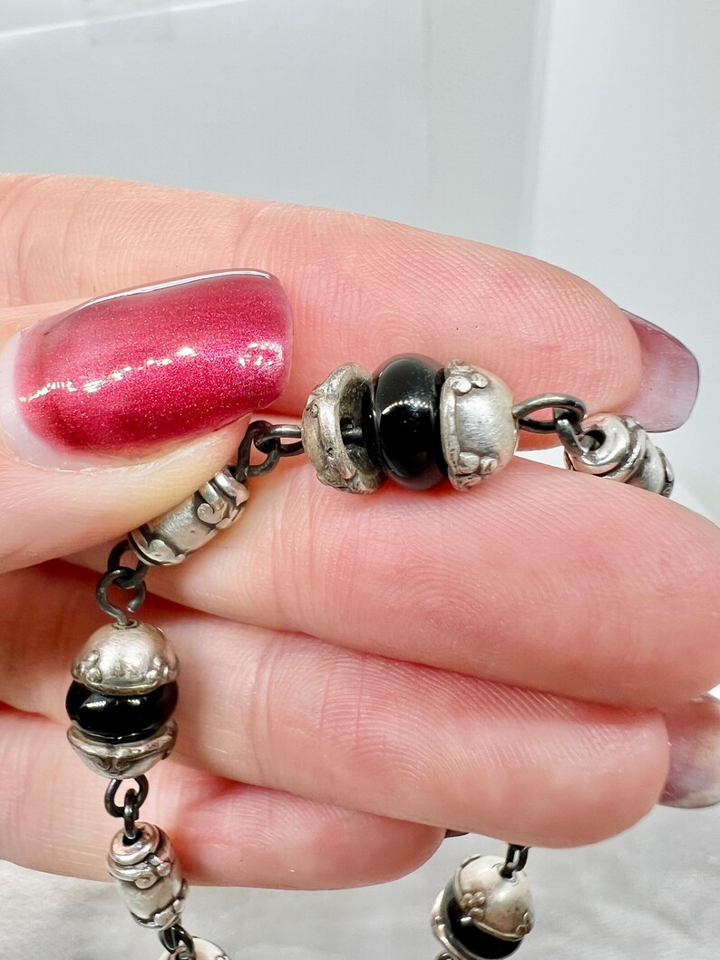 Puede incluir: Un brazalete de cadena de plata con cuentas negras y dijes de plata. El brazalete est&aacute; hecho de una cadena delicada y presenta una variedad de dijes, incluyendo peque&ntilde;as flores y otros elementos decorativos.