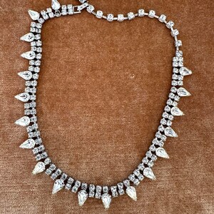 May include: A silver-toned necklace with clear, sparkling rhinestones. The necklace features a double-strand design with teardrop-shaped stones evenly spaced along the bottom strand. The necklace is displayed on a brown surface.