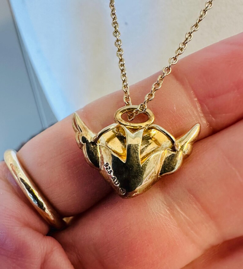 K&ouml;nnte beinhalten: Eine goldfarbene Halskette mit einem herzf&ouml;rmigen Anh&auml;nger mit Fl&uuml;geln. Der Anh&auml;nger hat ge&auml;tzte Details und eine filigrane Kette. Die Halskette ist mit "925 AU" markiert.