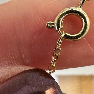 K&ouml;nnte beinhalten: Nahaufnahme eines goldfarbenen Halskettenverschlusses und einer Kette. Der Verschluss ist kreisf&ouml;rmig mit einem kleinen Hebel. Eine dunkelviolette, ovale Perle ist teilweise sichtbar, was auf eine Perlenkette hindeutet. Kette und Verschluss scheinen aus Gold zu sein.