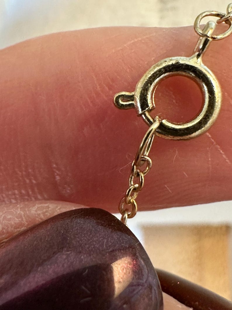 K&ouml;nnte beinhalten: Nahaufnahme eines goldfarbenen Halskettenverschlusses und einer Kette. Der Verschluss ist kreisf&ouml;rmig mit einem kleinen Hebel. Eine dunkelviolette, ovale Perle ist teilweise sichtbar, was auf eine Perlenkette hindeutet. Kette und Verschluss scheinen aus Gold zu sein.