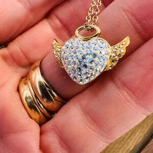 K&ouml;nnte beinhalten: Eine goldfarbene Halskette mit einem herzf&ouml;rmigen Anh&auml;nger, der mit Kristallen und kleinen goldenen Fl&uuml;geln verziert ist. Der Anh&auml;nger h&auml;ngt an einer zarten Goldkette. Die Halskette wird in einer Hand gehalten, mit goldenen Ringen an den Fingern.