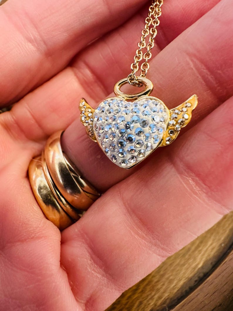 K&ouml;nnte beinhalten: Eine goldfarbene Halskette mit einem herzf&ouml;rmigen Anh&auml;nger, der mit Kristallen und kleinen goldenen Fl&uuml;geln verziert ist. Der Anh&auml;nger h&auml;ngt an einer zarten Goldkette. Die Halskette wird in einer Hand gehalten, mit goldenen Ringen an den Fingern.