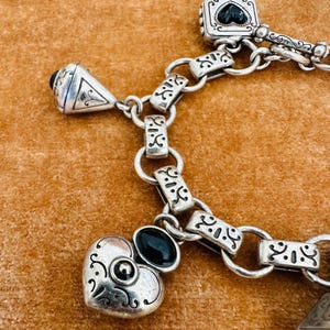 May include: Close-up of a silver bracelet with ornate detailing. The bracelet features various charms, including a heart, a square with a black heart, and a triangular charm. The bracelet is connected by a chain with decorative links. The charms have black accents.