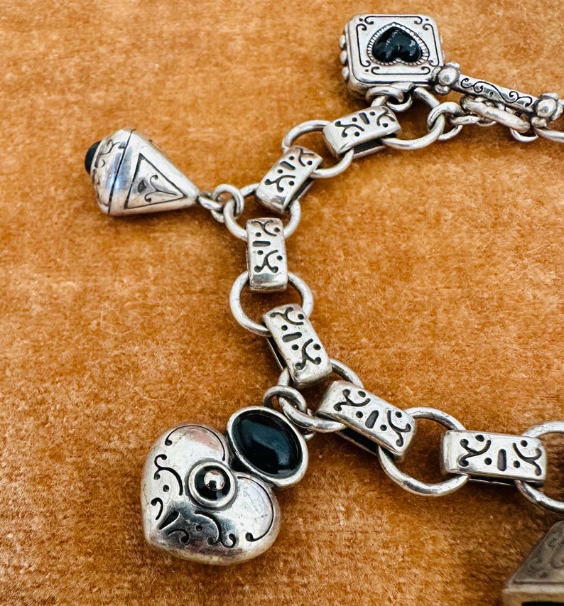 May include: Close-up of a silver bracelet with ornate detailing. The bracelet features various charms, including a heart, a square with a black heart, and a triangular charm. The bracelet is connected by a chain with decorative links. The charms have black accents.